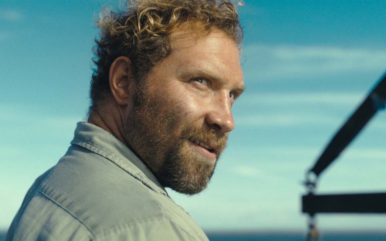 Bärtiger Mann mit lockigem Haar blickt an einem sonnigen Tag über die Schulter; im Hintergrund Meer und blauer Himmel.